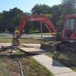 A red excavator in the front yard of a brick home in a forested area.