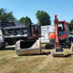 A man in shades with a red excavator and large truck performs an excavation in front of a home.