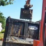 A red excavator and a crane behind a Professional Tank & Environmental Truck in a neighborhood.