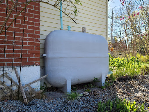 A new oil tank sits alongside a residential home. It is gray. The siding is beige in color with brick highlights. You can see part of the backyard.